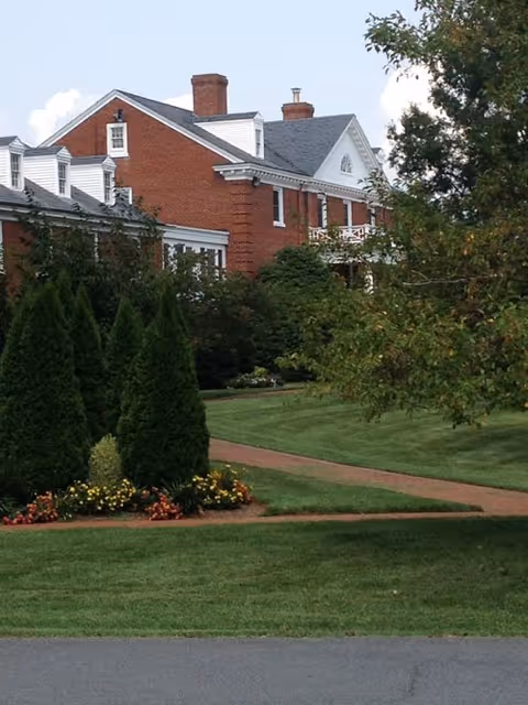A large brick building with white trim and multiple chimneys, surrounded by well-maintained green lawns, trees, and flower beds. A paved walkway curves through the grass in front of the building.