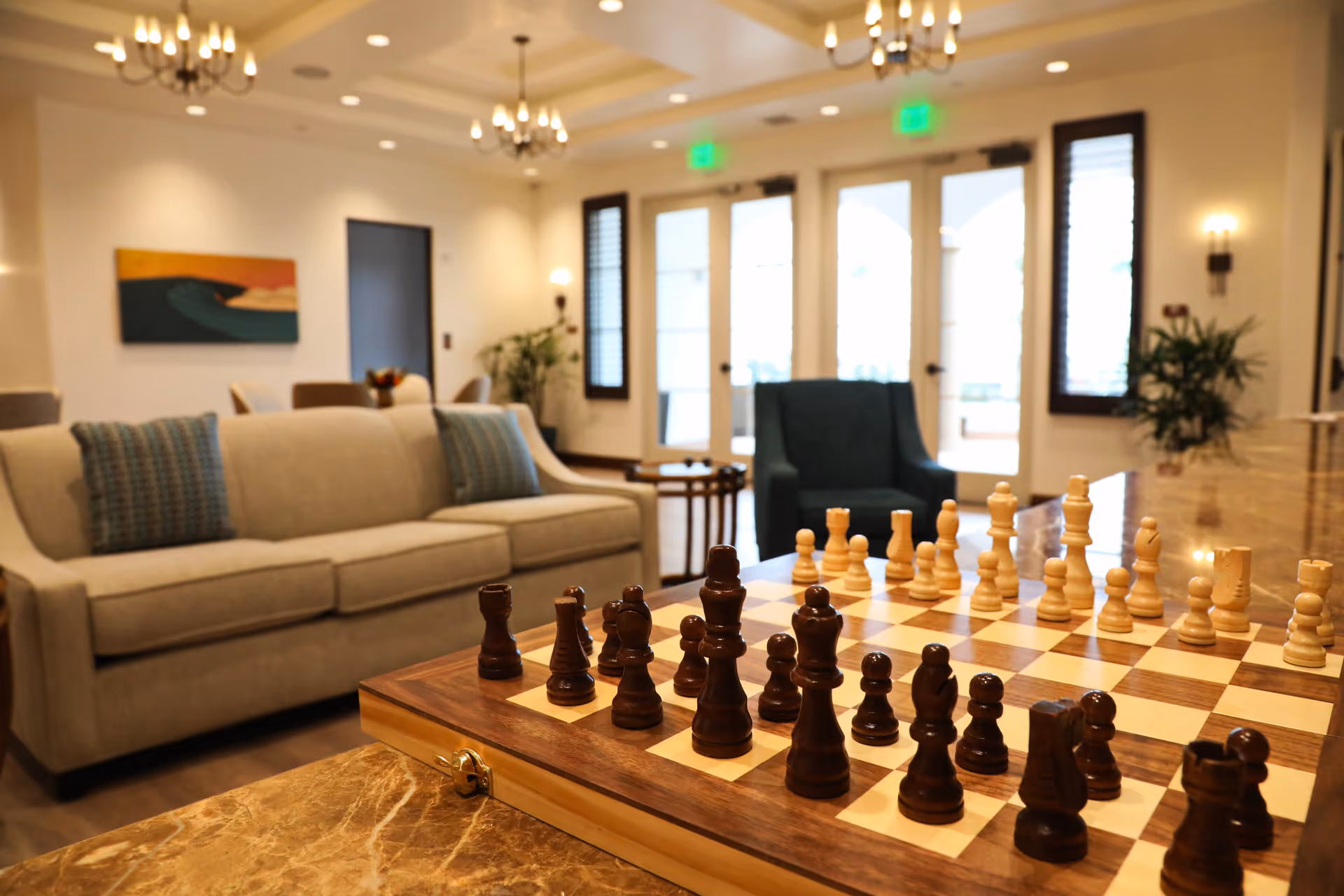 A cozy living room area with a beige sofa adorned with two blue patterned pillows, a dark blue armchair, and a wooden chessboard with chess pieces set up on a marble table in the foreground. The room has warm lighting from chandeliers and wall sconces, large windows with shutters, and a colorful painting on the wall.