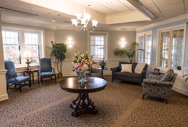 A cozy living room area with a round wooden table in the center holding a vase of flowers. The room has patterned carpet, two blue upholstered chairs near a window with a small table and lamp between them, a dark gray loveseat with two pillows, and a patterned armchair. There are plants near the windows and soft lighting from wall sconces and a chandelier.