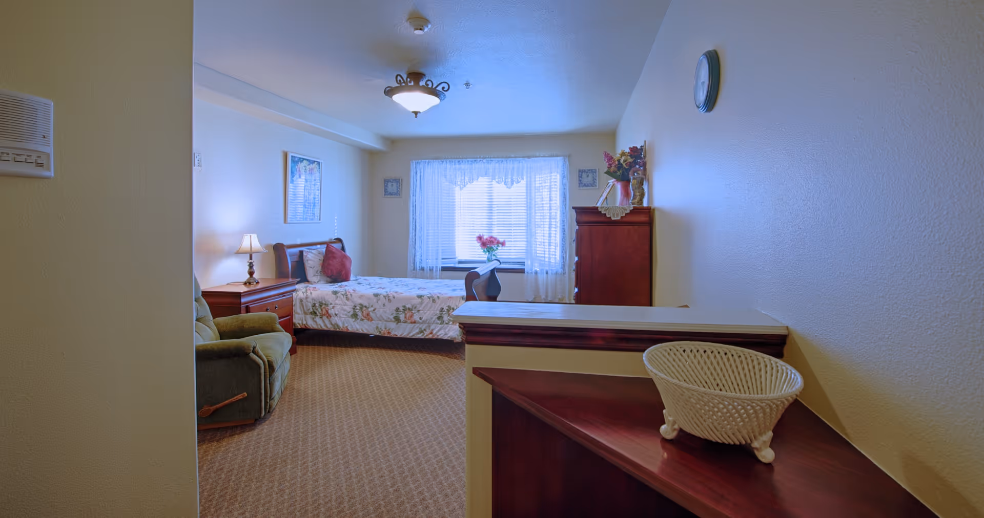 A cozy bedroom in a senior living facility with a single bed covered in floral bedding, a green recliner chair, a wooden nightstand with a lamp, a wooden dresser with decorative items, and a window with lace curtains letting in natural light.