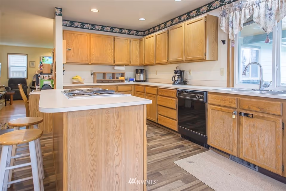 Bright kitchen with wood cabinets, a center island with cooktop and bar stools, dishwasher, and sink under a window.