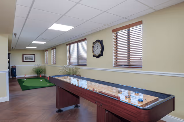Indoor recreational room with a shuffleboard table in the foreground and a small putting green with golf clubs in the background. The room has yellow walls, wooden blinds on the windows, a wall clock, and wood-patterned flooring.