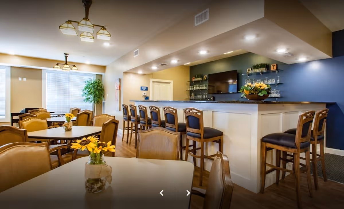 Interior view of a senior living facility dining and bar area with tables and chairs. The room features a long bar counter with high chairs, a mounted flat-screen TV, shelves with glassware, and decorative flower arrangements on the tables. Large windows allow natural light to brighten the space.