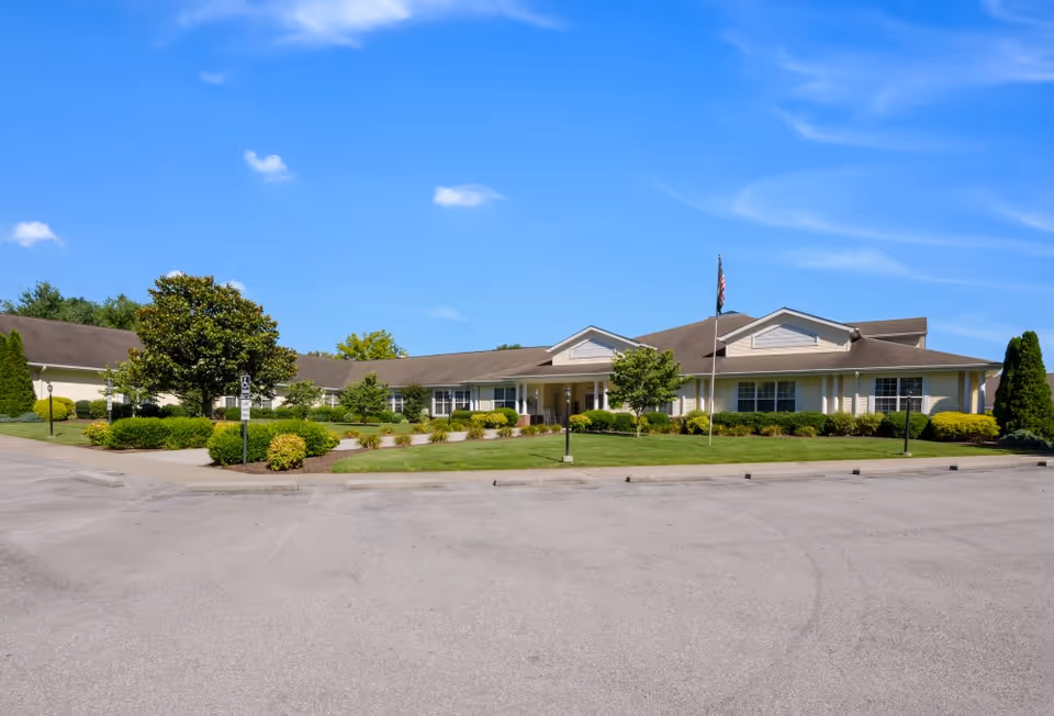 Exterior view of a single-story senior living facility building with a well-maintained lawn, shrubs, and trees under a clear blue sky. An American flag is displayed on a flagpole near the entrance.