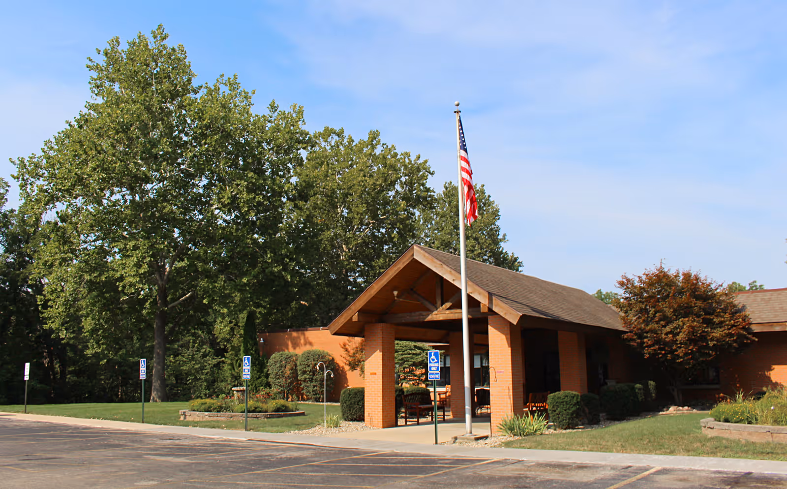 Exterior view of Mount Zion Health and Rehab Center showing the entrance with a covered drop-off area supported by brick columns. An American flag is flying on a flagpole in front of the entrance. There are trees, shrubs, and a well-maintained lawn surrounding the building under a clear blue sky.