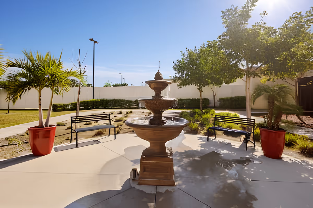 Outdoor courtyard area with a central tiered water fountain, two black metal benches on either side, and large potted plants in red containers. The area is surrounded by trees, shrubs, and a white fence under a clear blue sky with sunlight.
