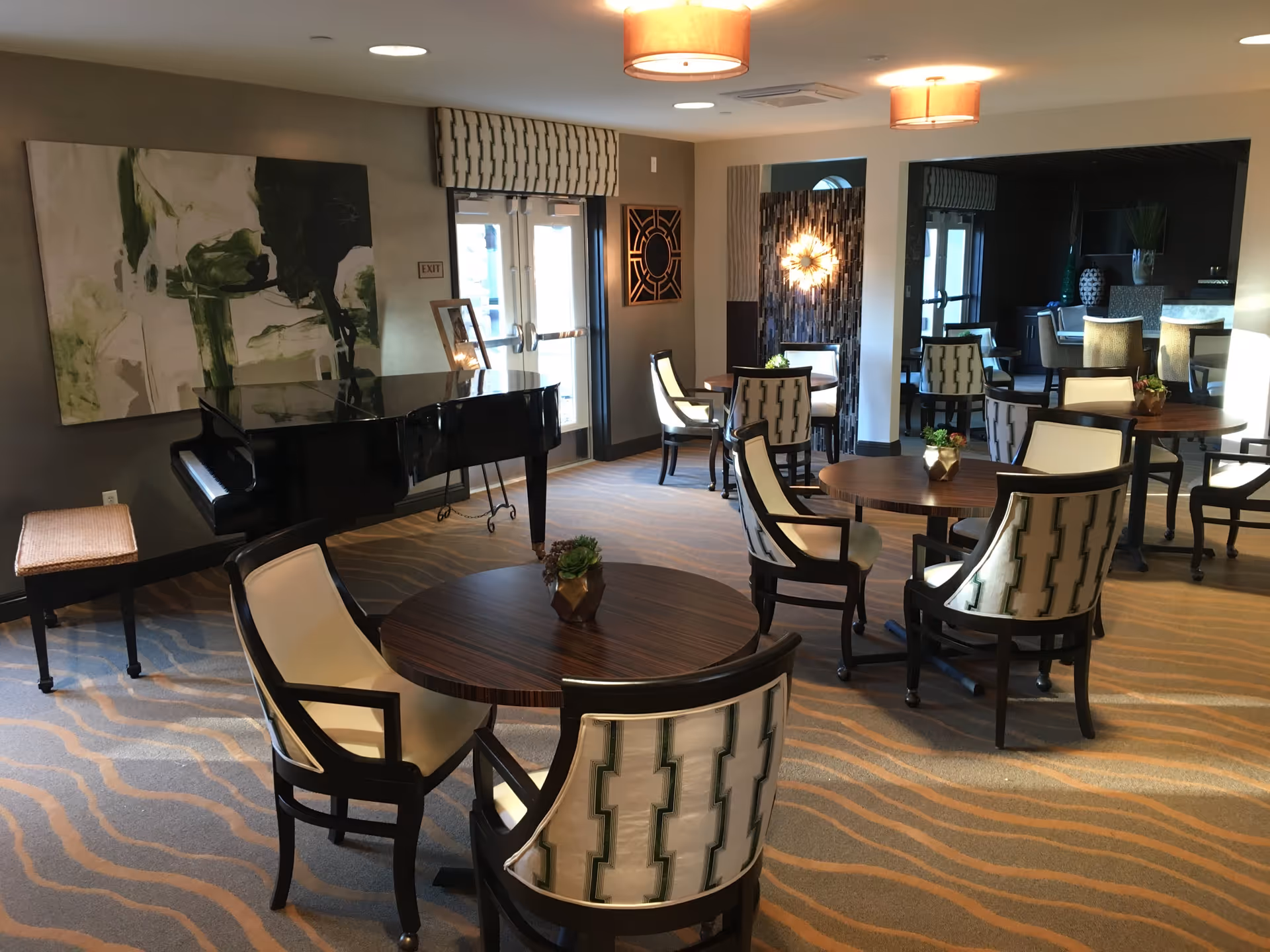 A well-lit common area with several round wooden tables surrounded by upholstered chairs with patterned backs. A black grand piano with a bench is positioned near a wall with abstract green and white artwork. The room has carpeted flooring with a wavy pattern, ceiling lights, and large windows allowing natural light to enter.