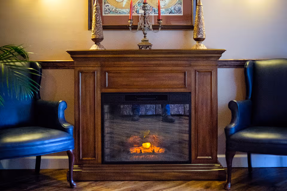 A cozy interior scene featuring a wooden electric fireplace with a glowing fire effect, flanked by two dark blue leather armchairs. On top of the fireplace mantel are two ornate candle holders with red candles and a decorative centerpiece. A framed artwork hangs on the wall above the mantel.