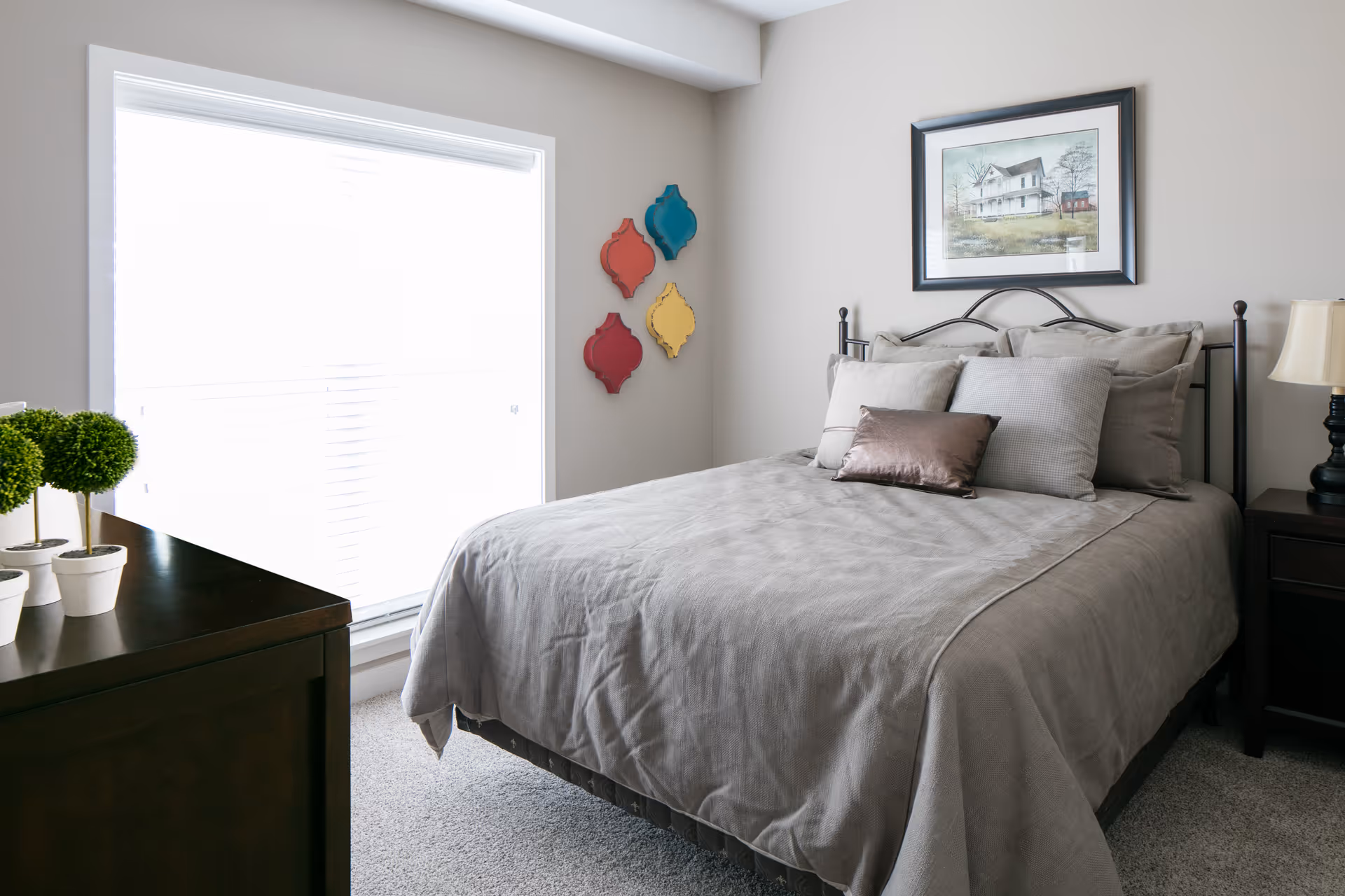 A neatly made bed in a bright bedroom with a dresser, nightstand, wall art and small potted plants by a large window.