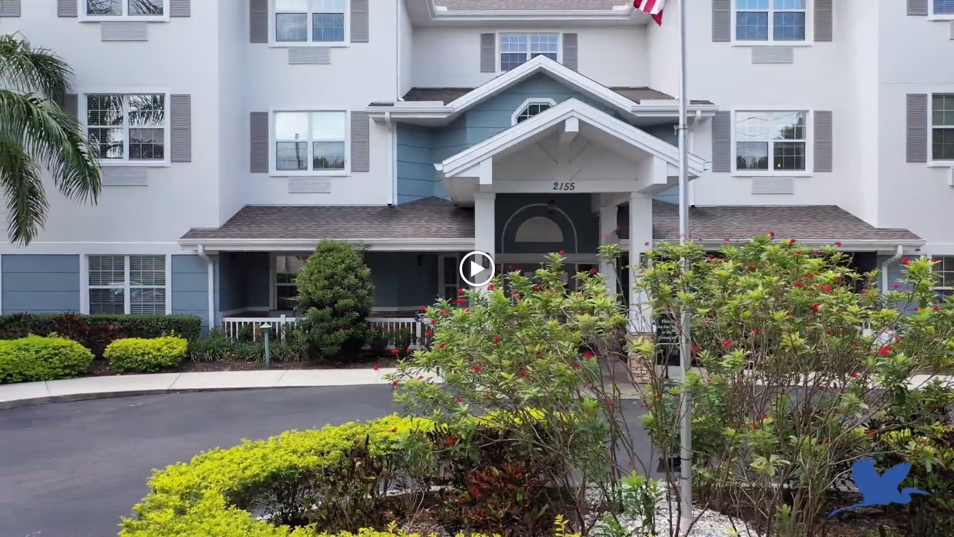 Front exterior view of a multi-story senior living facility building with white and blue siding, multiple windows with shutters, a covered entrance with the number 2155 above the door, an American flag on a flagpole, and landscaped bushes and greenery in front.