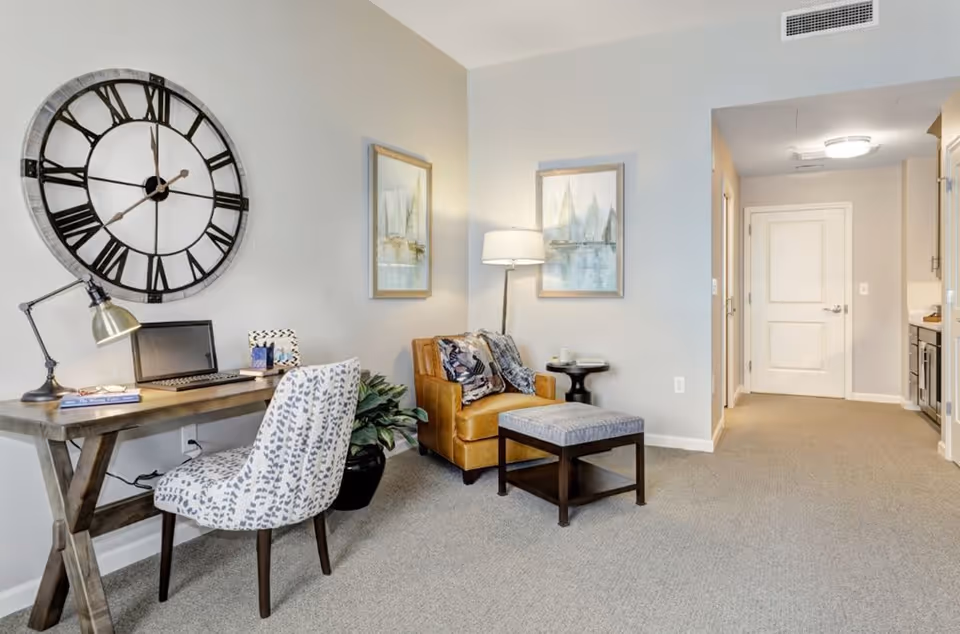 A cozy interior space featuring a wooden desk with a laptop, a patterned chair, a large wall clock, and a potted plant. Next to the desk is a comfortable leather armchair with a patterned throw pillow and a matching ottoman. Two framed paintings hang on the wall above the armchair, and a floor lamp provides soft lighting. In the background, there is a hallway leading to a white door and a glimpse of a kitchen area.