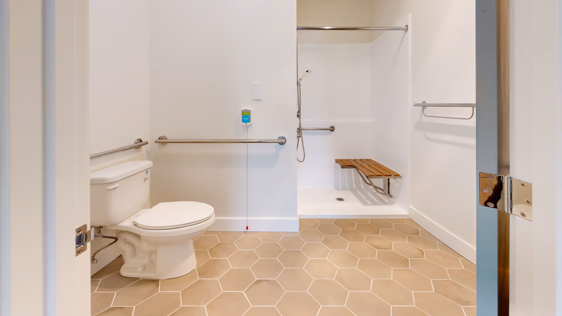 A clean, accessible bathroom with a white toilet, grab bars on the walls, and a walk-in shower with a foldable wooden bench and handheld showerhead. The floor has beige hexagonal tiles and the walls are painted white.