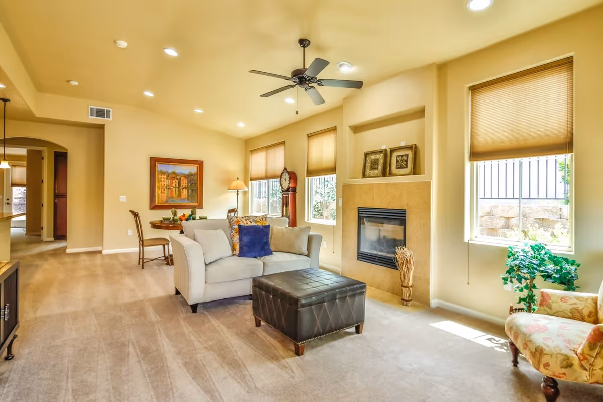 Bright and spacious living room with beige walls and carpeted floor. The room features a white sofa with colorful cushions, a dark brown leather ottoman, a floral armchair, a fireplace with decorative frames above it, and large windows with beige blinds letting in natural light. A ceiling fan with lights is mounted on the ceiling. In the background, there is a small dining area with a round table and chairs, a floor lamp, and a framed painting on the wall.