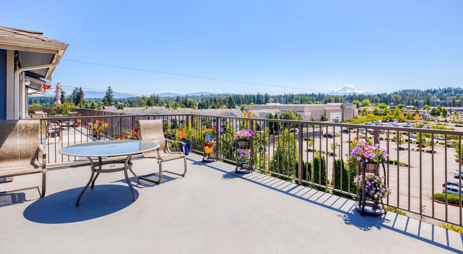 A spacious outdoor balcony with a glass-top round table and several chairs. The balcony has a metal railing adorned with colorful flower pots. In the background, there is a view of a parking lot, trees, buildings including a Kohl's store, and distant mountains under a clear blue sky.