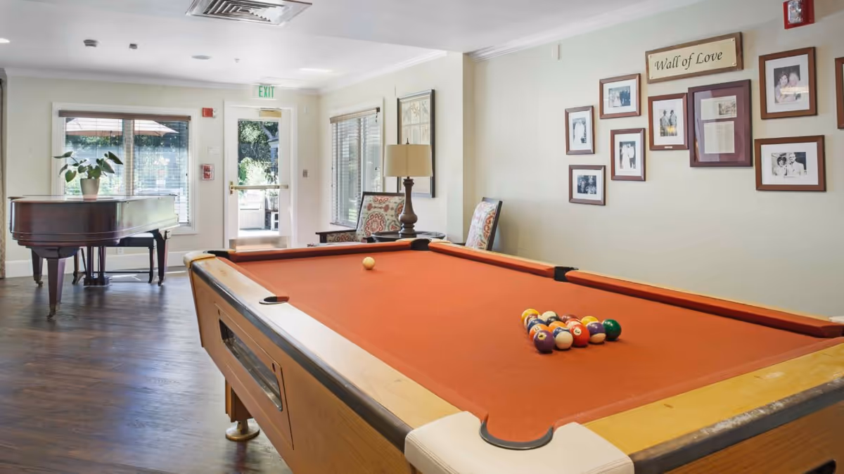 A bright room with a pool table featuring a red felt surface and a rack of billiard balls arranged for a game. In the background, there is a grand piano with a potted plant on top, two patterned armchairs, a floor lamp, and a wall decorated with framed photos and a sign that reads 'Wall of Love'. Large windows and a glass door let in natural light, showing an outdoor patio area.