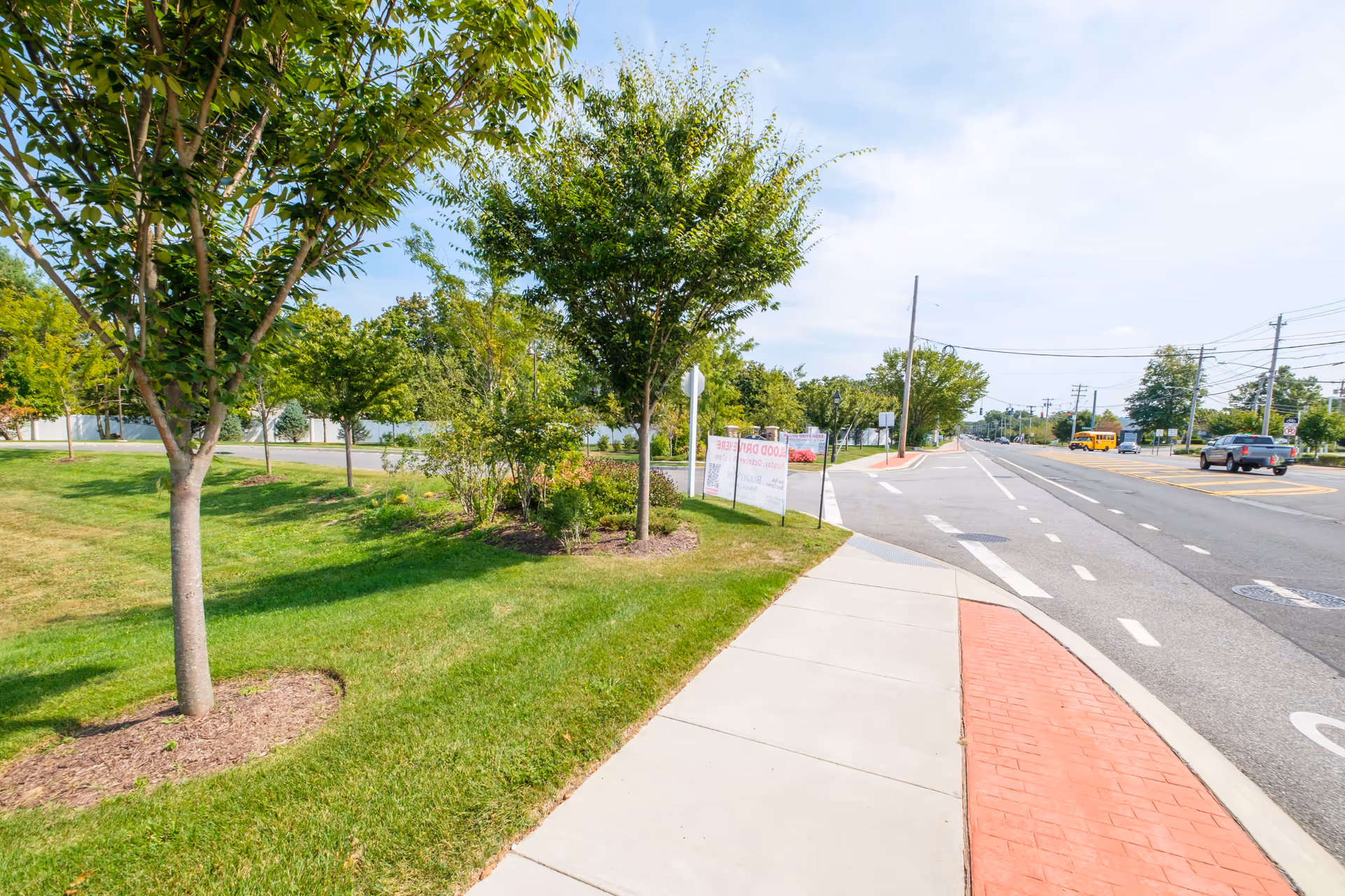A sidewalk runs alongside a grassy area with several young trees and shrubs. Across the street, there are vehicles including a yellow school bus and a pickup truck. The sky is partly cloudy and the scene appears to be in a suburban area.