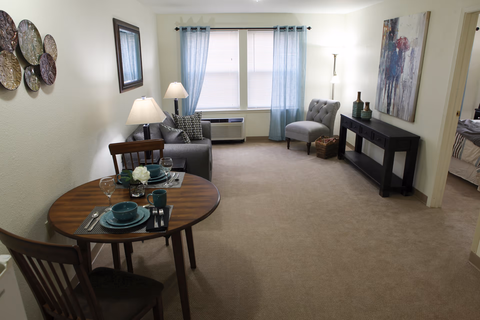 A cozy living room area with a small wooden dining table set for two with teal dishes and glassware. There is a gray sofa with patterned pillows, a gray upholstered chair, and a black console table with decorative vases and a colorful abstract painting above it. Light blue curtains frame a window with blinds, and the room is softly lit by two lamps.
