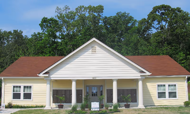 Front exterior view of a single-story building with a brown roof, white columns, and yellow siding. The building has a central entrance door flanked by multiple mailboxes and potted plants. Trees are visible in the background under a clear blue sky.