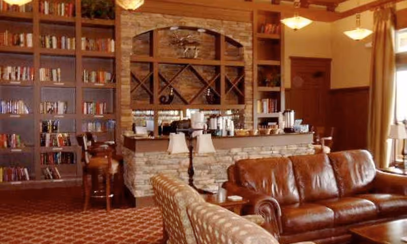 A cozy living room area with a brown leather sofa and a patterned armchair facing a stone bar counter. Behind the bar, there are shelves with decorative items and a built-in wine rack. To the left, there is a large bookshelf filled with books. The room features warm lighting and wood accents.