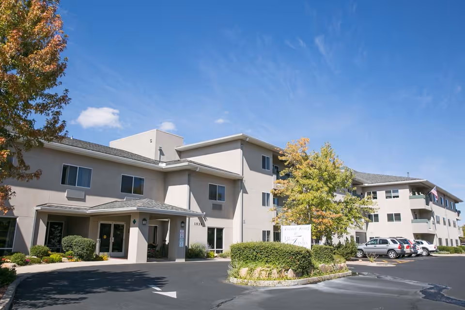 Exterior view of a three-story senior living facility building with beige walls and multiple windows. There is a covered entrance with a driveway and several parked cars. Trees with green and autumn-colored leaves are visible around the building under a clear blue sky.