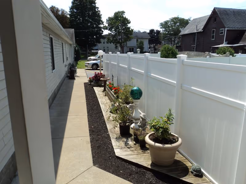 Narrow outdoor side walkway beside a building lined with potted plants and a white vinyl fence.