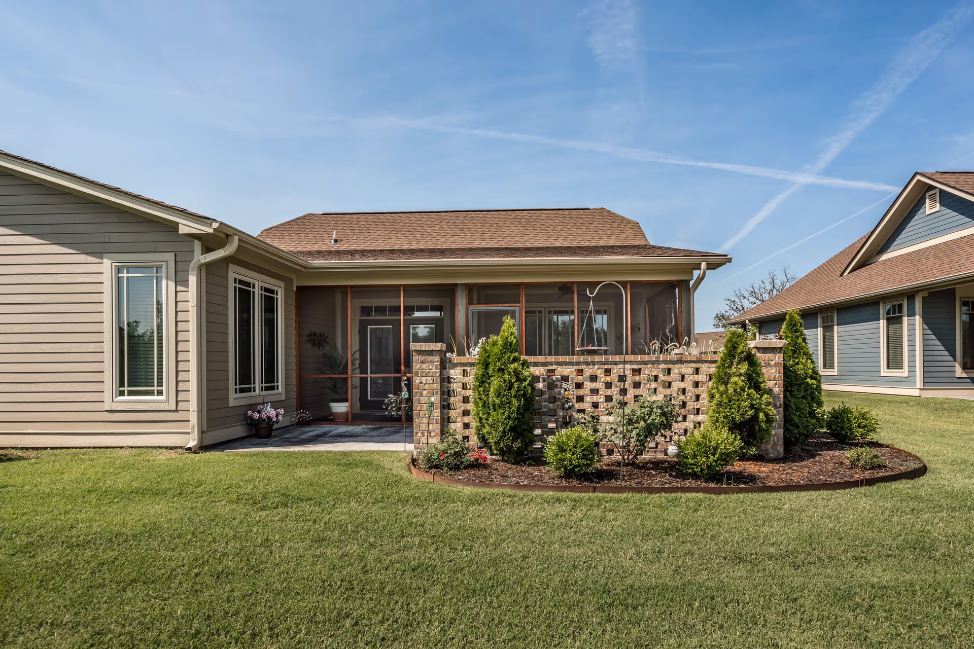 Exterior view of a single-story residential building with beige siding and a brown roof. The building features a screened porch area with a brick privacy wall and landscaping including small bushes and flowers in front. Another similar building is visible in the background under a clear blue sky.