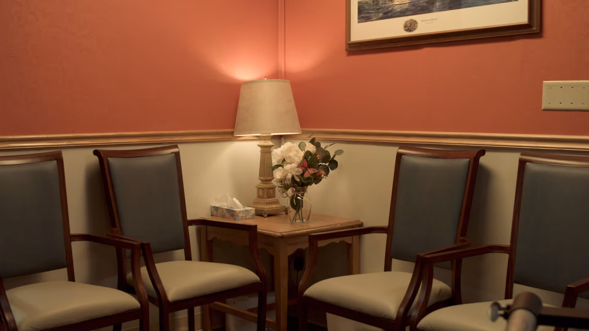 Upholstered wooden chairs arranged around a small side table with a lamp, tissue box, and vase of flowers in a softly lit waiting area.
