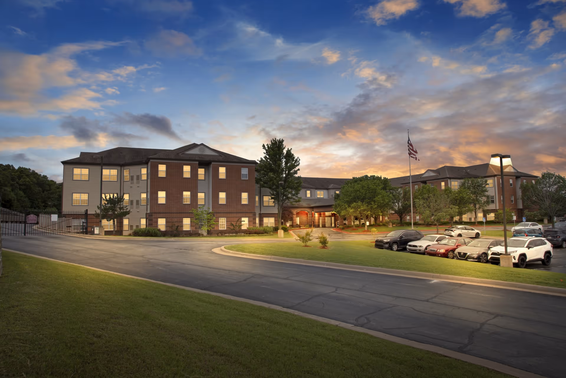 Exterior front of a three-story senior living building at dusk with lit windows, a parking lot, and an American flag.