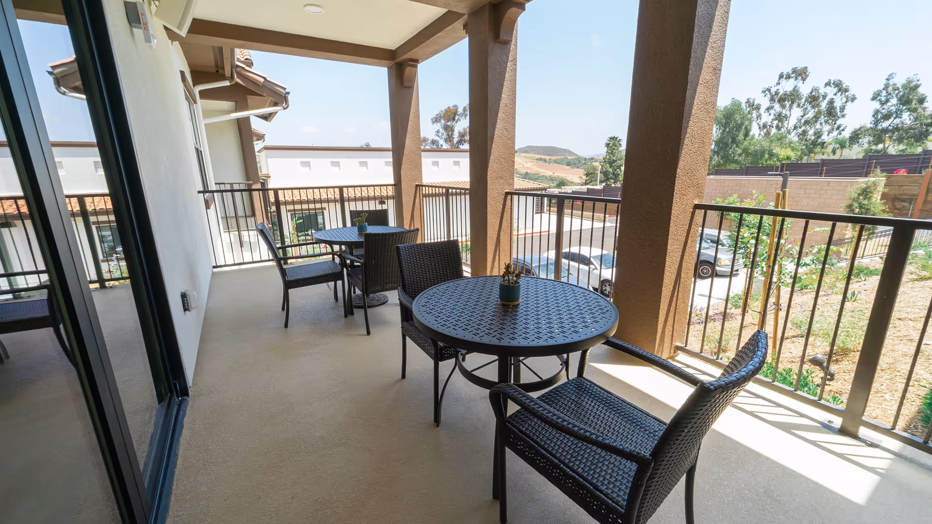 Outdoor covered balcony area with two round black metal tables, each accompanied by two black woven chairs. Small potted plants are placed on each table. The balcony overlooks a parking area with cars and some greenery, with hills visible in the distance under a clear sky.