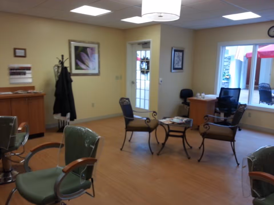 Bright assisted-living salon/common room with styling chairs, a small seating area and a desk near a window.
