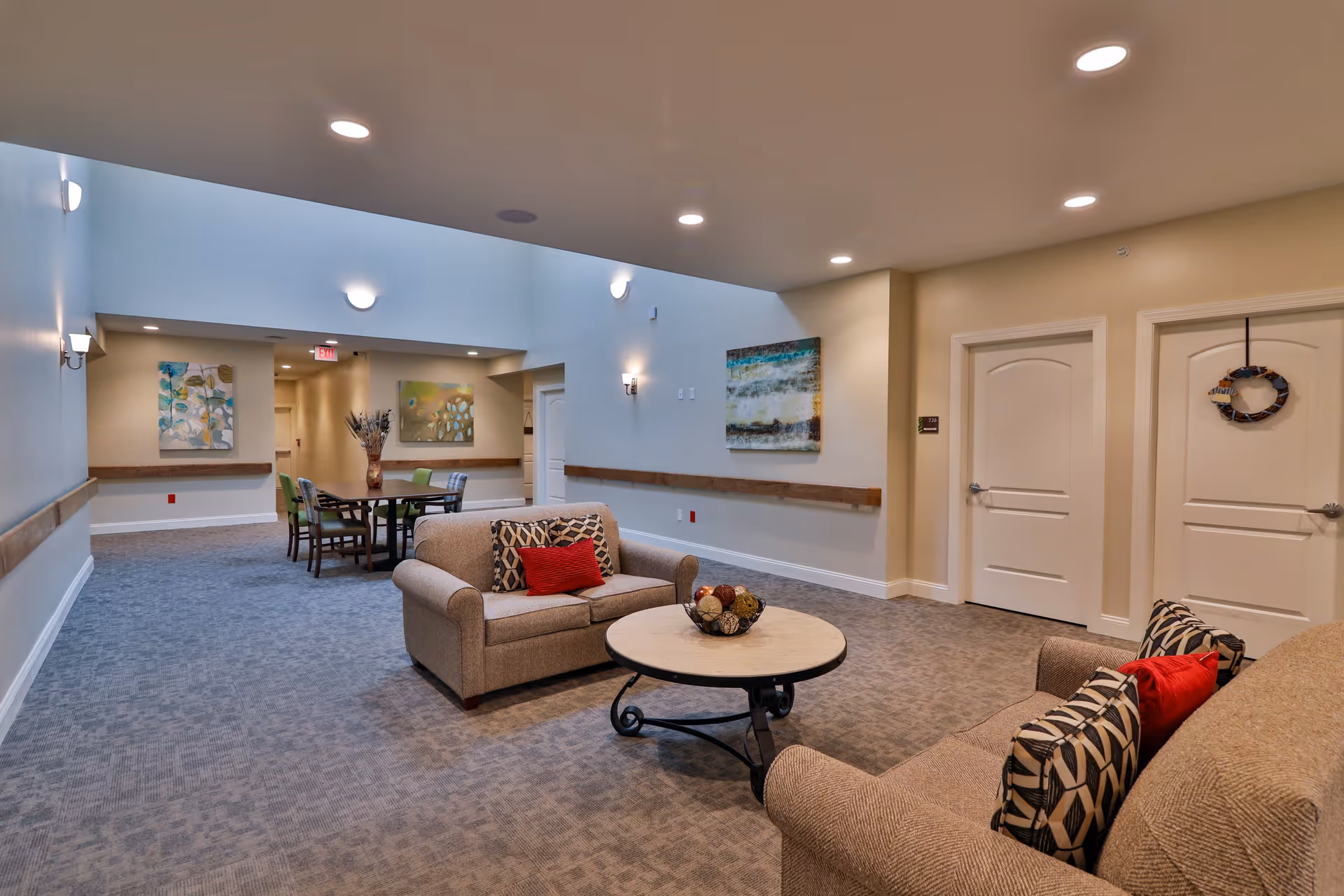 A spacious and well-lit common area in a senior living facility featuring two beige sofas with patterned and red cushions, a round coffee table with decorative items, and a dining table with chairs in the background. The walls are adorned with colorful abstract paintings, and there are multiple doors and soft lighting fixtures on the ceiling and walls.