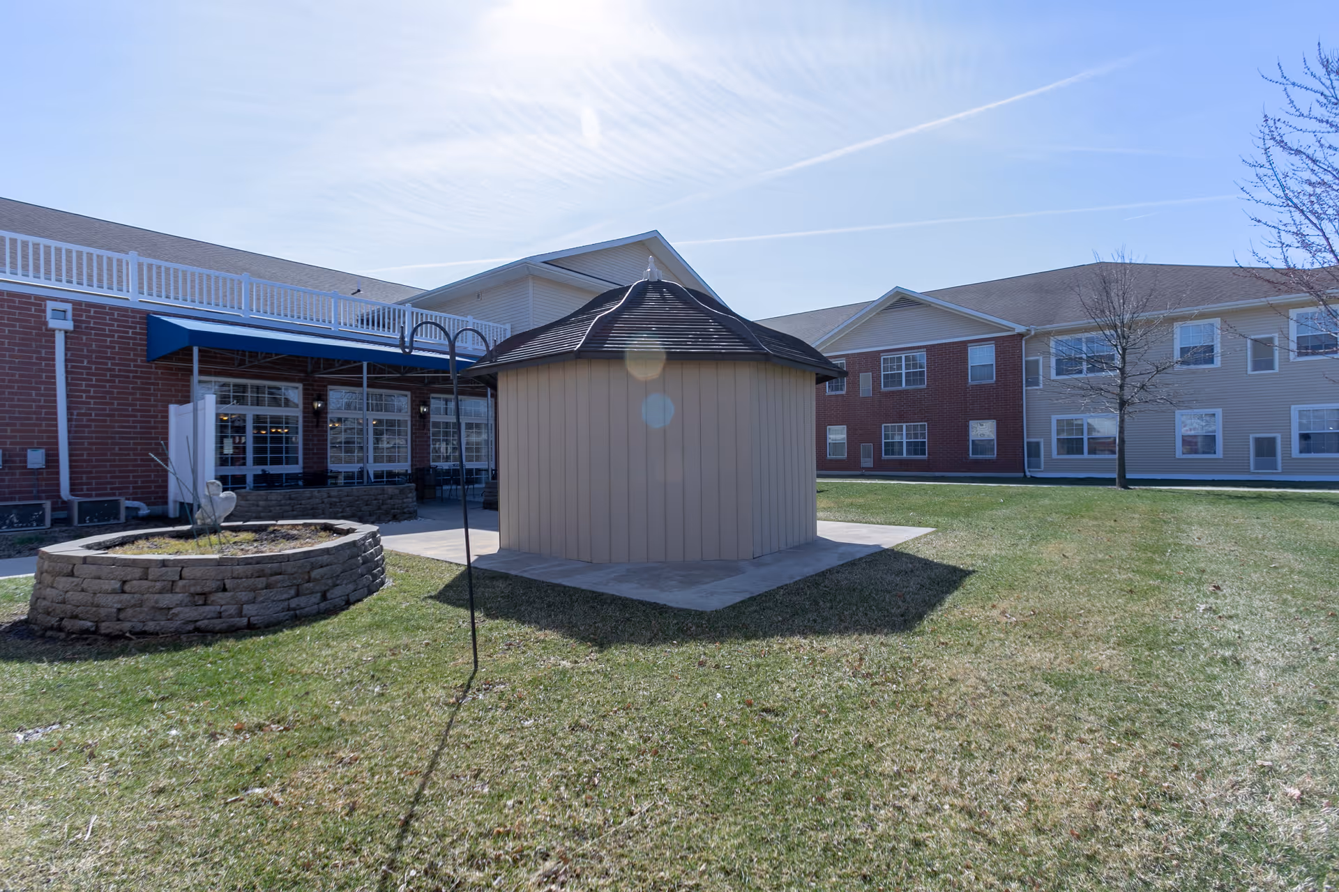 A small octagonal gazebo sits on a grassy courtyard in front of a two-story senior living building with a stone planter and a blue awning.