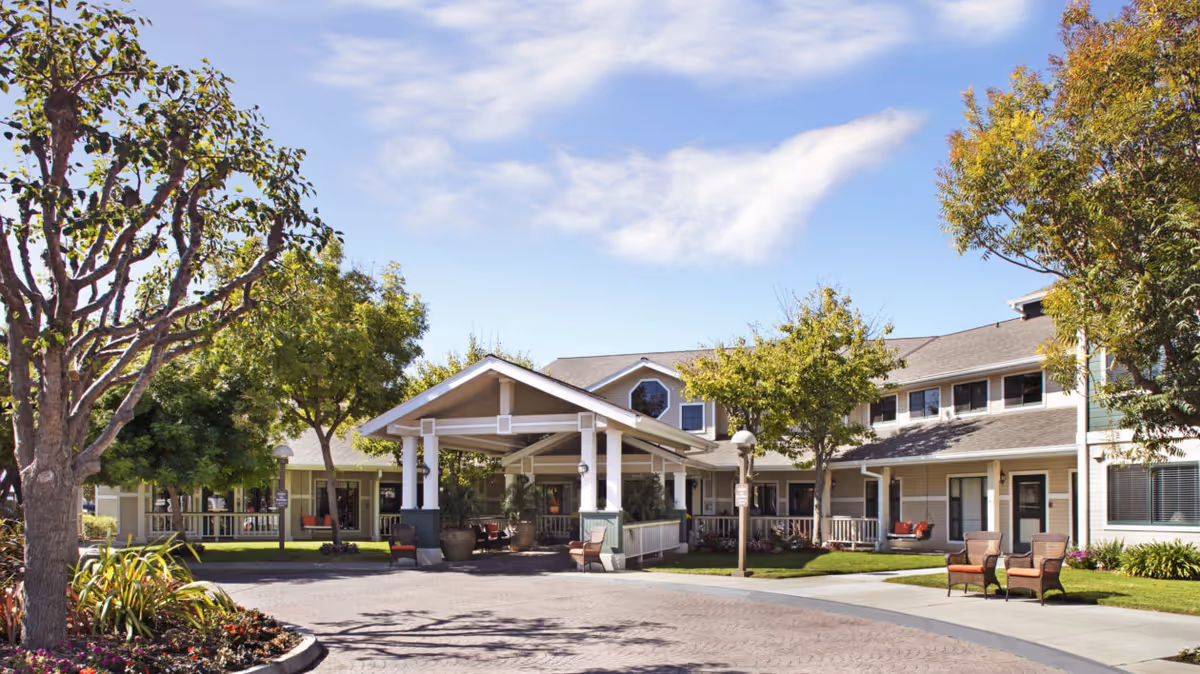 Exterior view of a senior living facility with a covered entrance, surrounded by trees and landscaped greenery under a blue sky with some clouds.