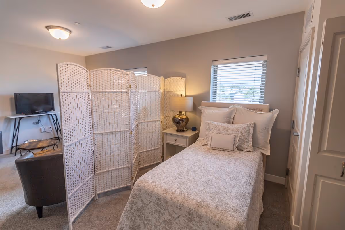 A cozy bedroom with a single bed covered in a light patterned bedspread and multiple pillows. Next to the bed is a white nightstand with a decorative lamp. A white folding room divider stands beside the bed, partially separating the sleeping area from a small sitting area with a dark chair and a TV on a stand. The room has beige walls, carpeted floor, and two windows with blinds.