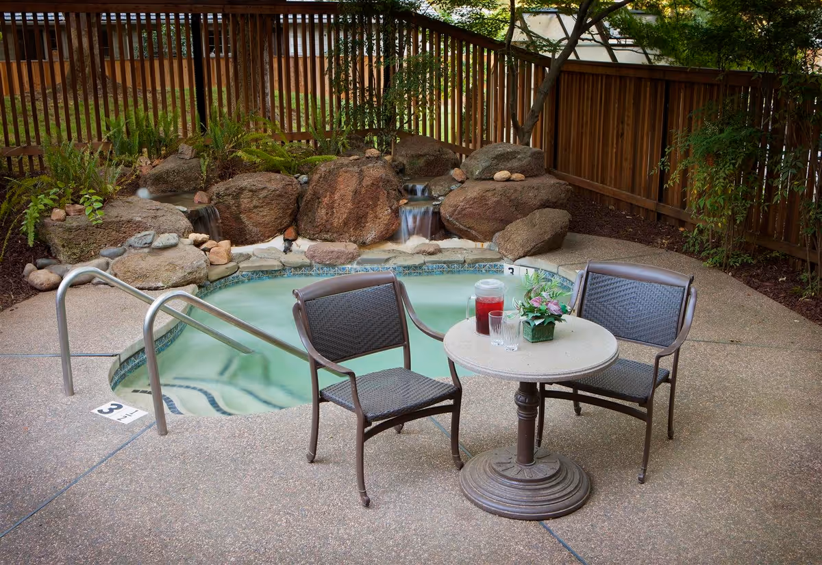 A small outdoor hot tub surrounded by rocks and plants with a wooden fence in the background. In front of the hot tub, there is a round table with two chairs. On the table, there is a pitcher of red beverage, two glasses, and a small flower arrangement.