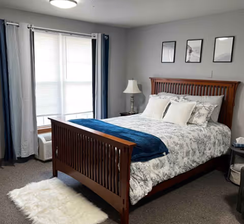 A neatly made bed with a wooden headboard and footboard in a bedroom. The bed has white and gray floral bedding with white pillows and a blue throw blanket. There is a white fluffy rug on the floor beside the bed. A window with white blinds and blue and white curtains is on the left side. A bedside table with a lamp and a small round table with a tissue box are visible. Three framed pictures hang on the light gray wall above the bed.