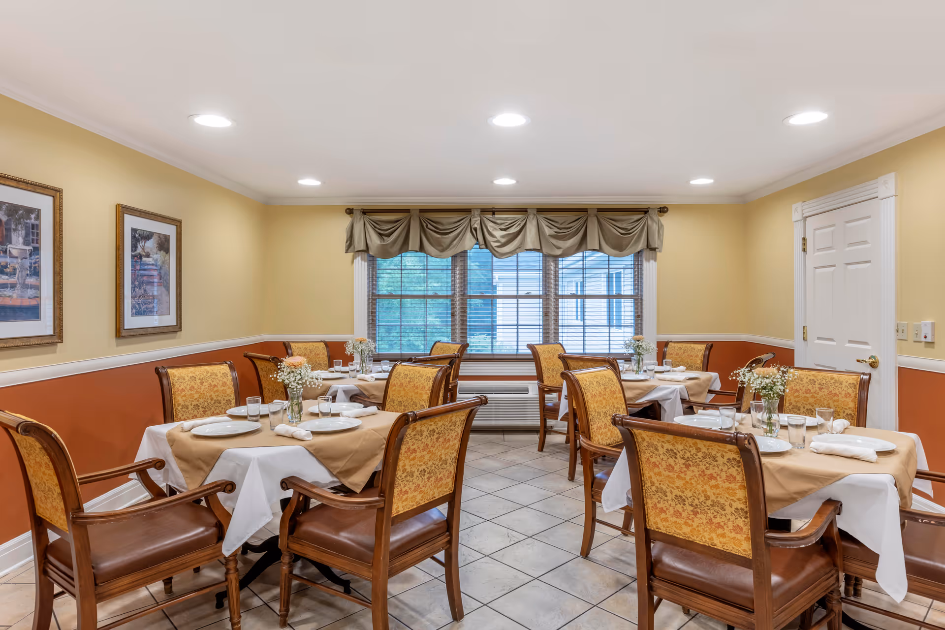 A cozy dining room with four tables, each set with white plates, glasses, and napkins. The tables have beige tablecloths with white underlays. The chairs have wooden frames with patterned upholstery. The room has warm yellow and orange walls, tile flooring, and a large window with beige curtains letting in natural light. Two framed pictures hang on the left wall.