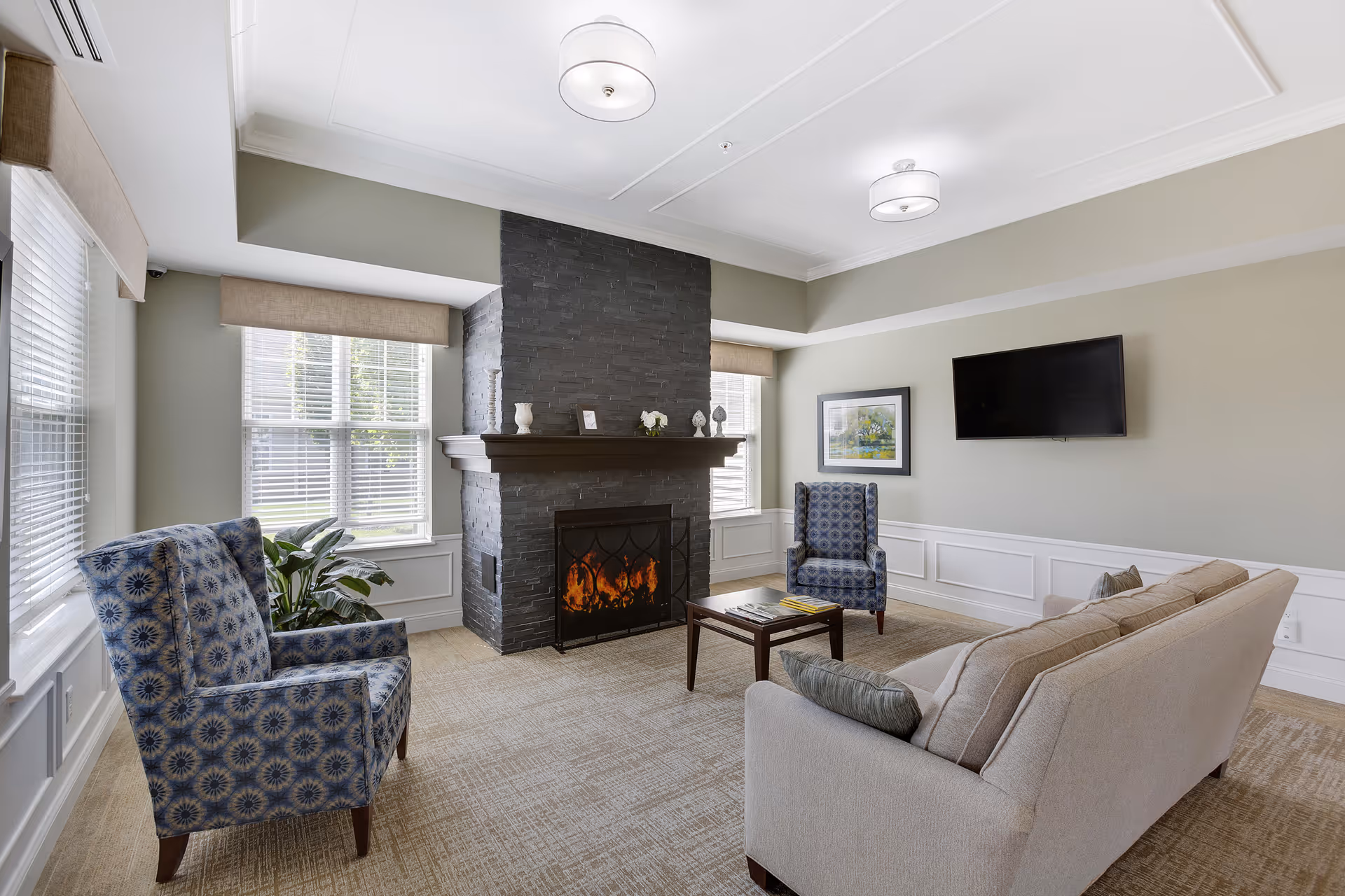 A cozy living room with a beige sofa, two blue patterned armchairs, a dark wooden coffee table, and a black stone fireplace with a fire burning. The room has large windows with blinds and beige valances, light gray walls with white wainscoting, a wall-mounted flat-screen TV, and two ceiling light fixtures.