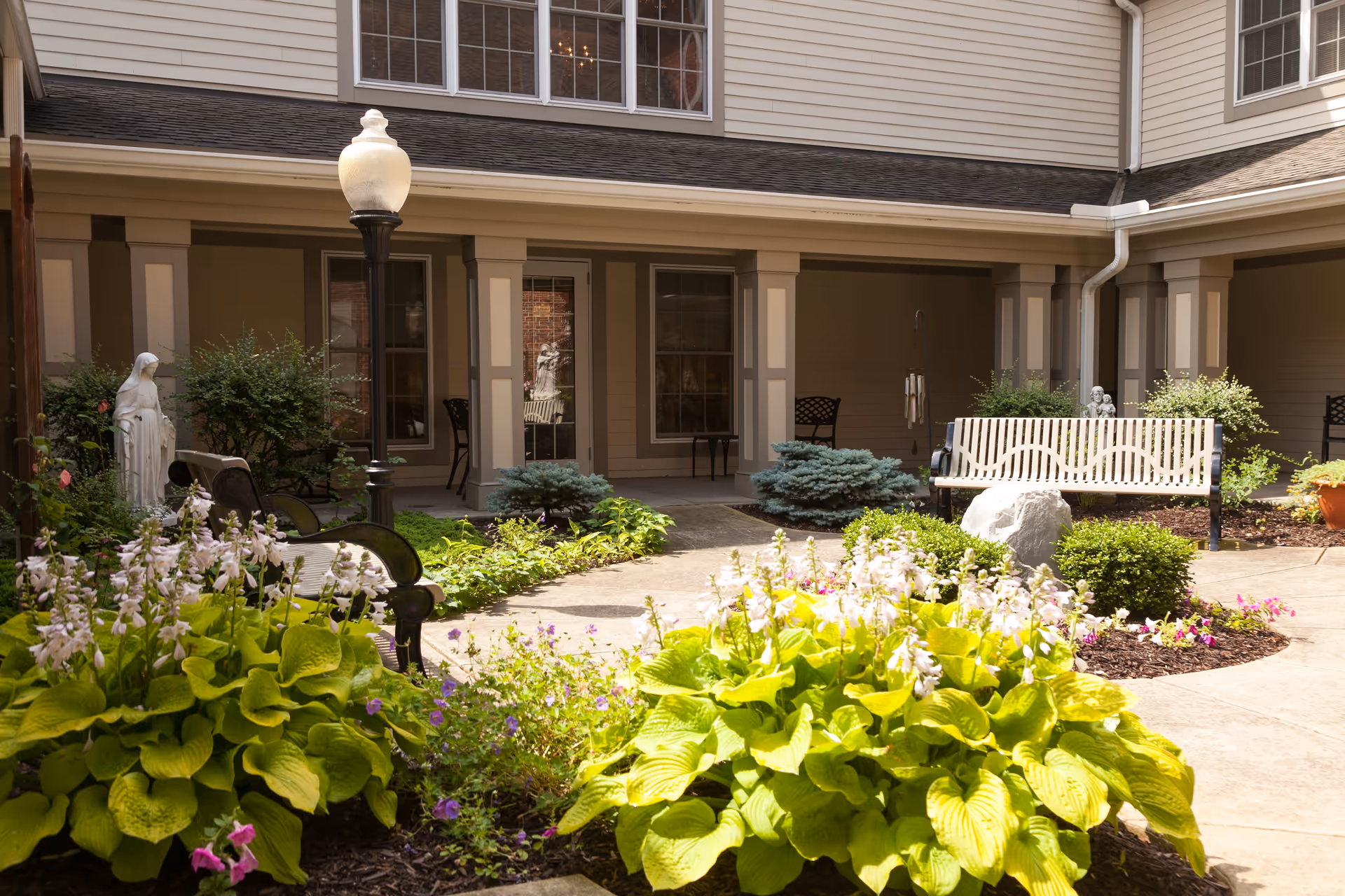 Sunlit courtyard with flowering beds, benches, a lamp post and a statue in front of a covered porch of a senior living building.