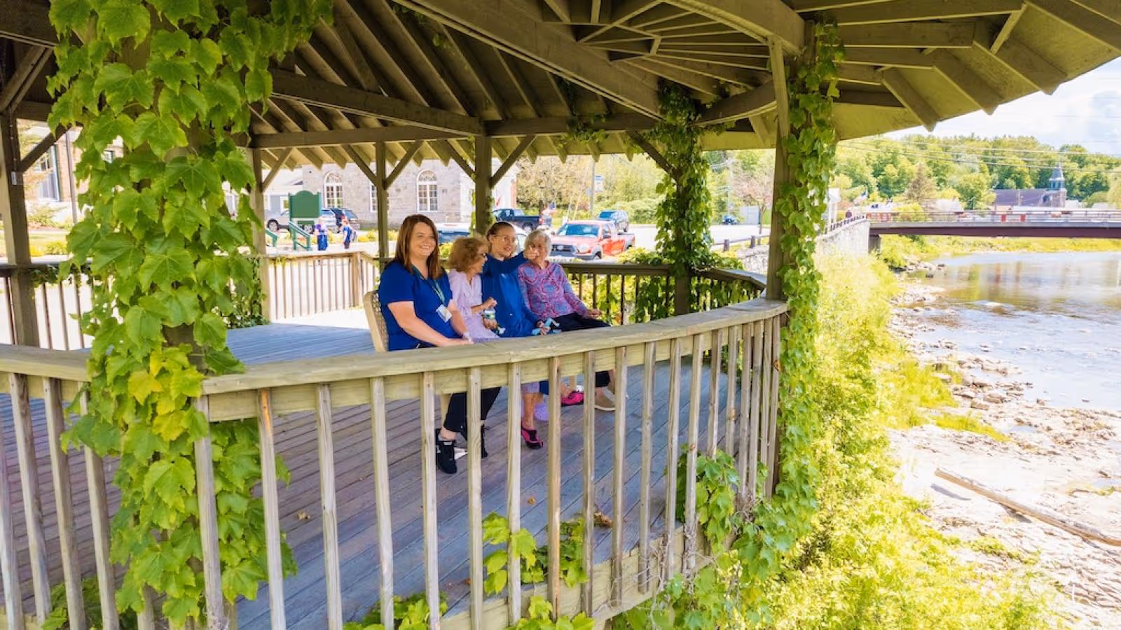 Four women sitting on a wooden bench under a gazebo covered with green vines, overlooking a river with a bridge in the background on a sunny day.