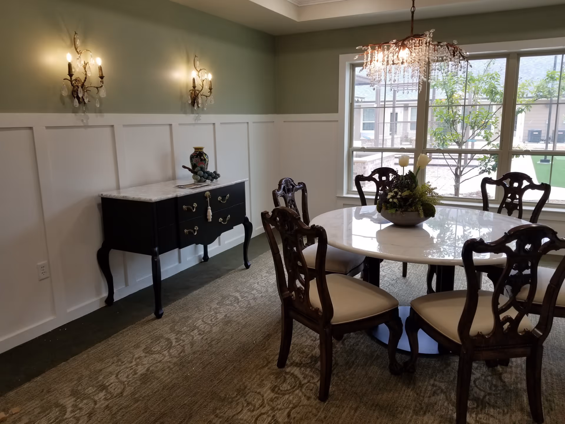 Elegant dining room with a round marble table surrounded by carved wooden chairs, a chandelier, sideboard, and large windows.