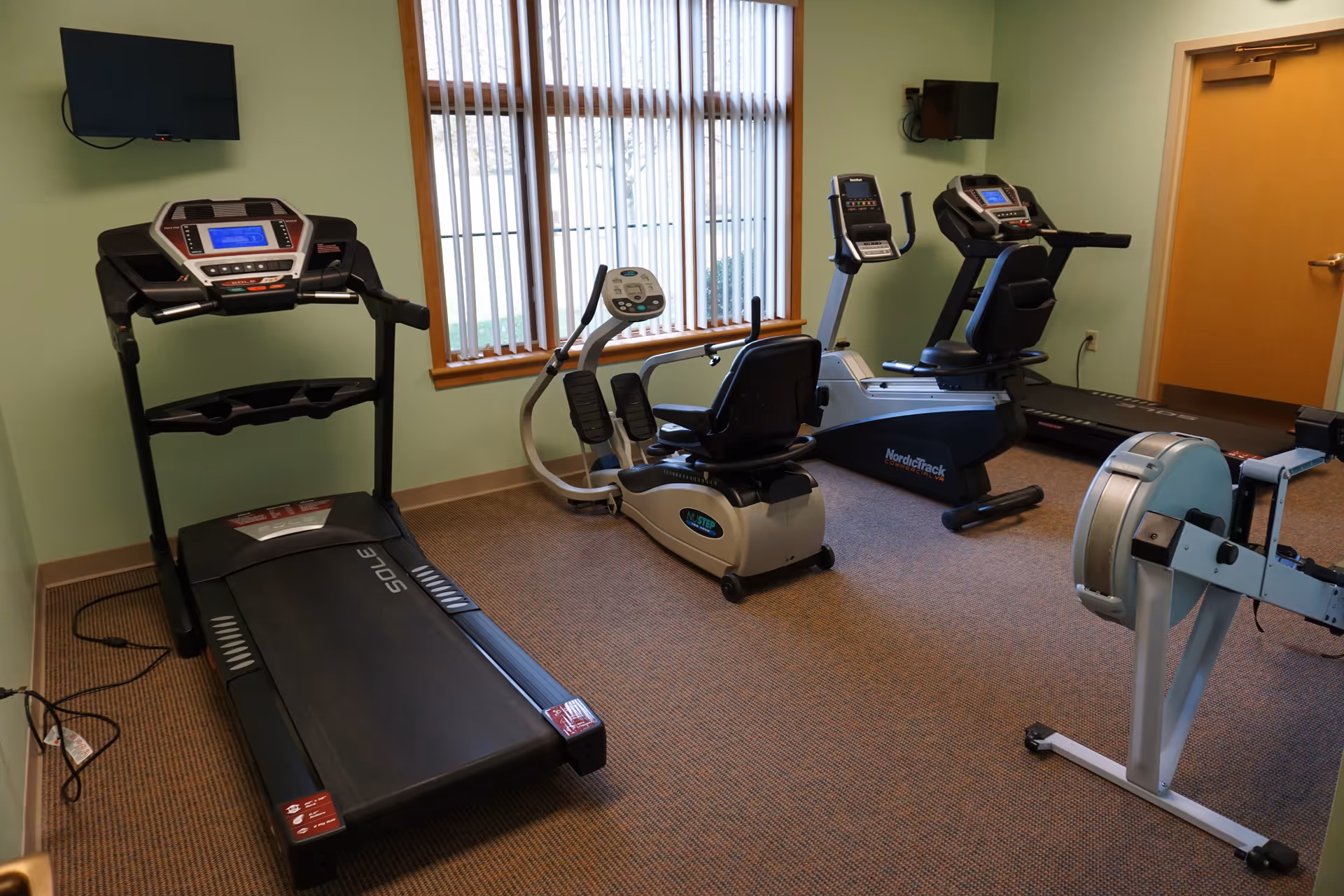 Small fitness room with treadmills, a recumbent bike, a rowing machine, and wall-mounted TVs by a window.