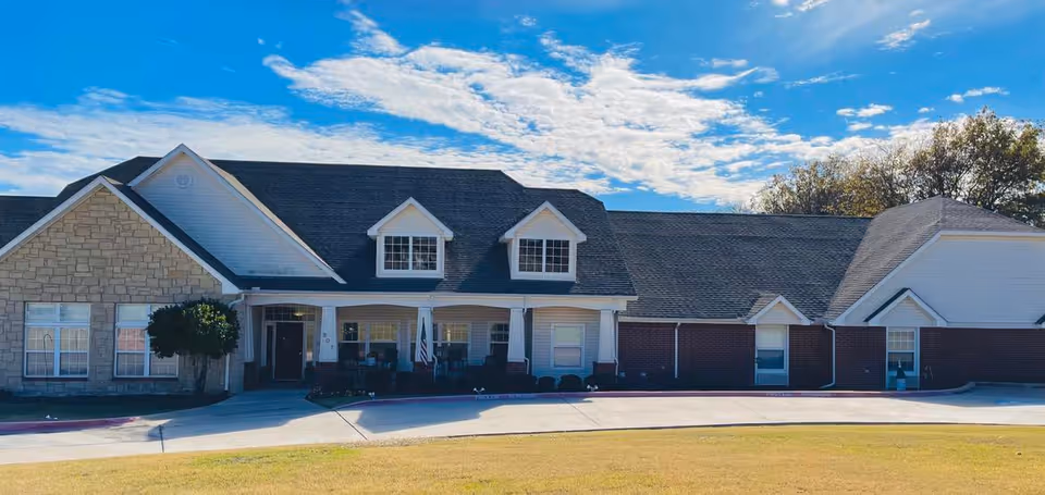 Front exterior view of Kyrie Assisted Living facility showing a single-story building with a combination of stone and brick facade, multiple windows, a covered porch with seating, and a driveway in front under a partly cloudy blue sky.