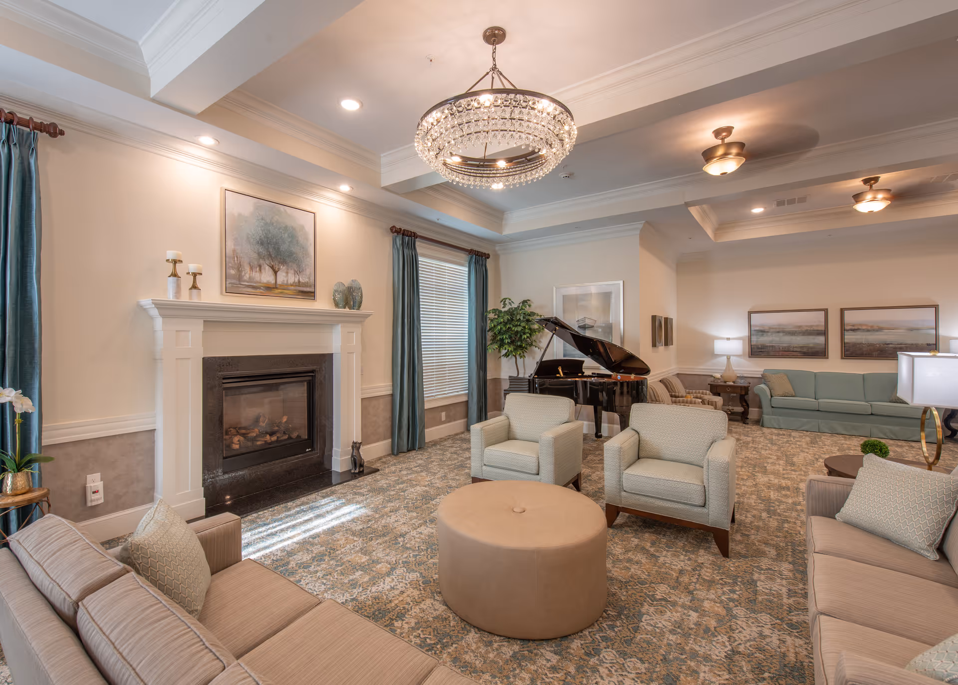 A spacious and elegant living room with beige sofas and armchairs arranged around a round ottoman. The room features a fireplace with decorative candles and artwork above it, teal curtains on the windows, a grand piano in the corner, and soft lighting from ceiling fixtures and lamps. The carpet has a patterned design, and the walls are painted in neutral tones.