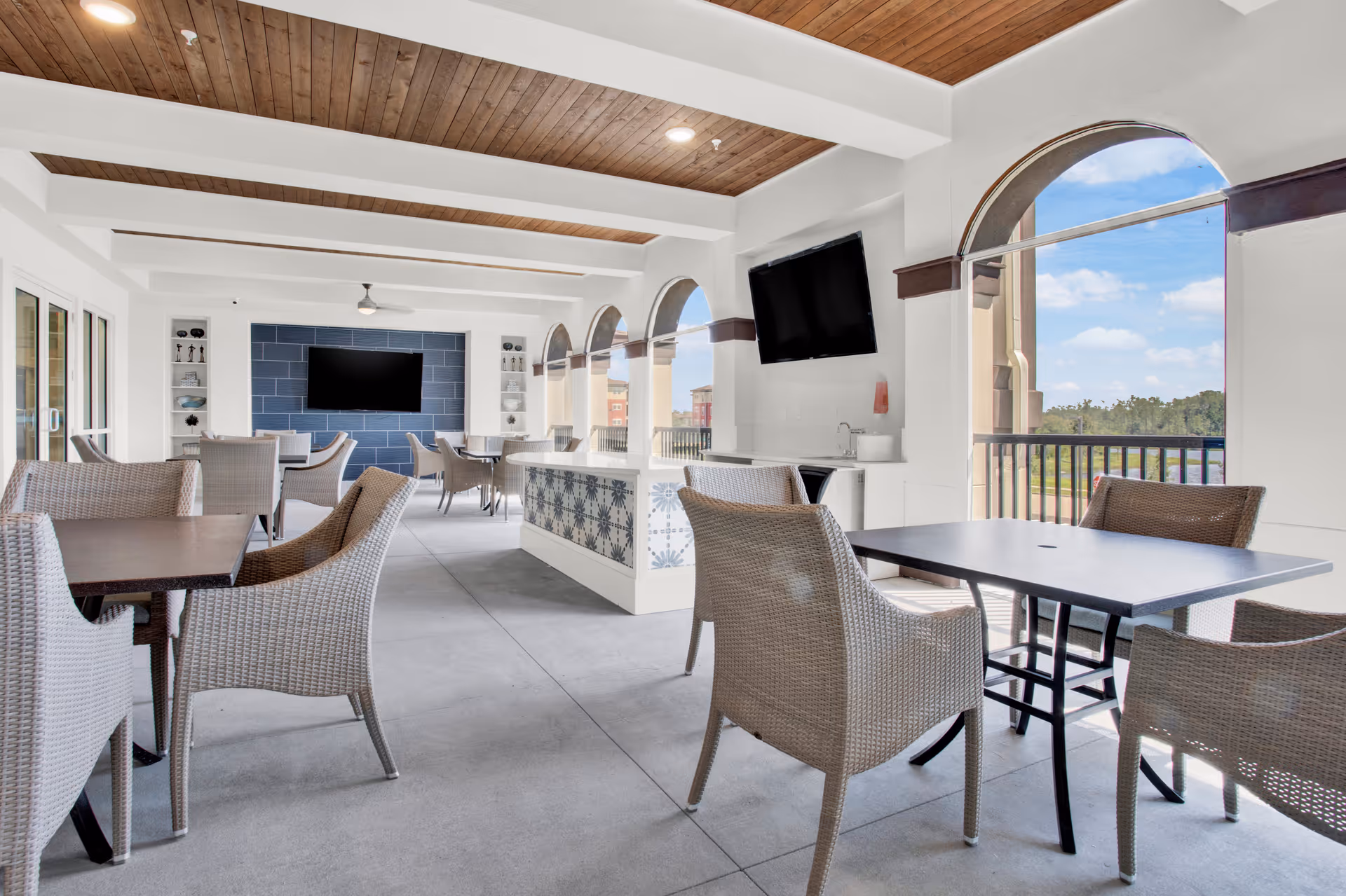 A bright and airy indoor lounge area with multiple tables and wicker chairs. The space features a wooden ceiling with white beams, large arched windows offering a view of the outdoors, two wall-mounted flat-screen TVs, and a decorative tiled counter in the center.