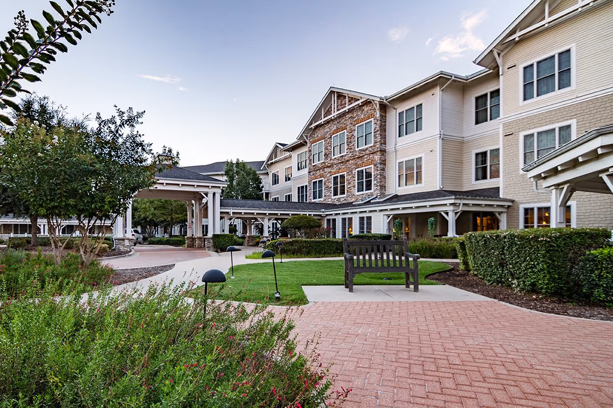 Exterior view of a multi-story senior living building with a landscaped courtyard, bench, and covered entrance.