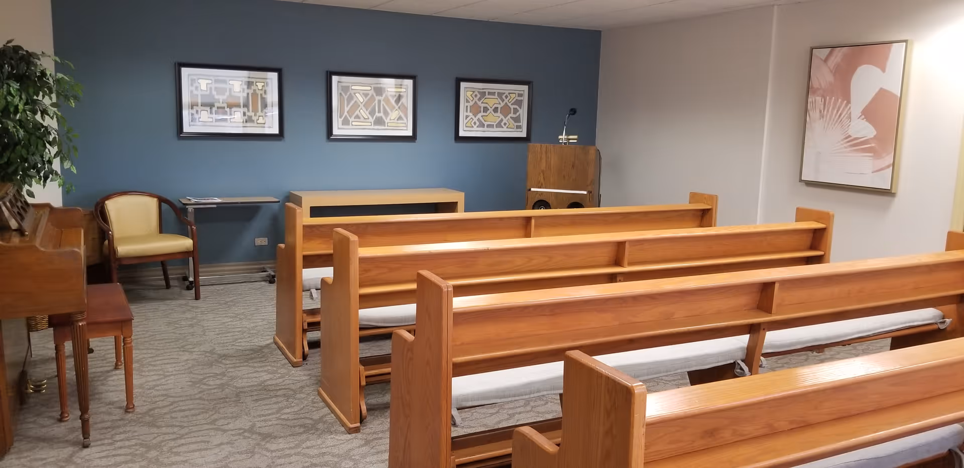 A small chapel or meditation room with wooden pews arranged in rows facing a small podium. The room has a blue accent wall with three framed abstract artworks, a piano with a bench on the left side, and a single chair next to a small table. The floor is carpeted and there is a framed abstract artwork on the right wall.