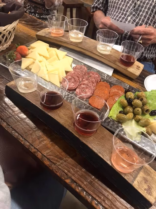 A wooden serving board on a rustic wooden table with an assortment of sliced cheeses, salami, pepperoni, green and black olives on lettuce, and several small plastic cups filled with different types of wine. Two people are partially visible in the background, one holding a piece of paper.