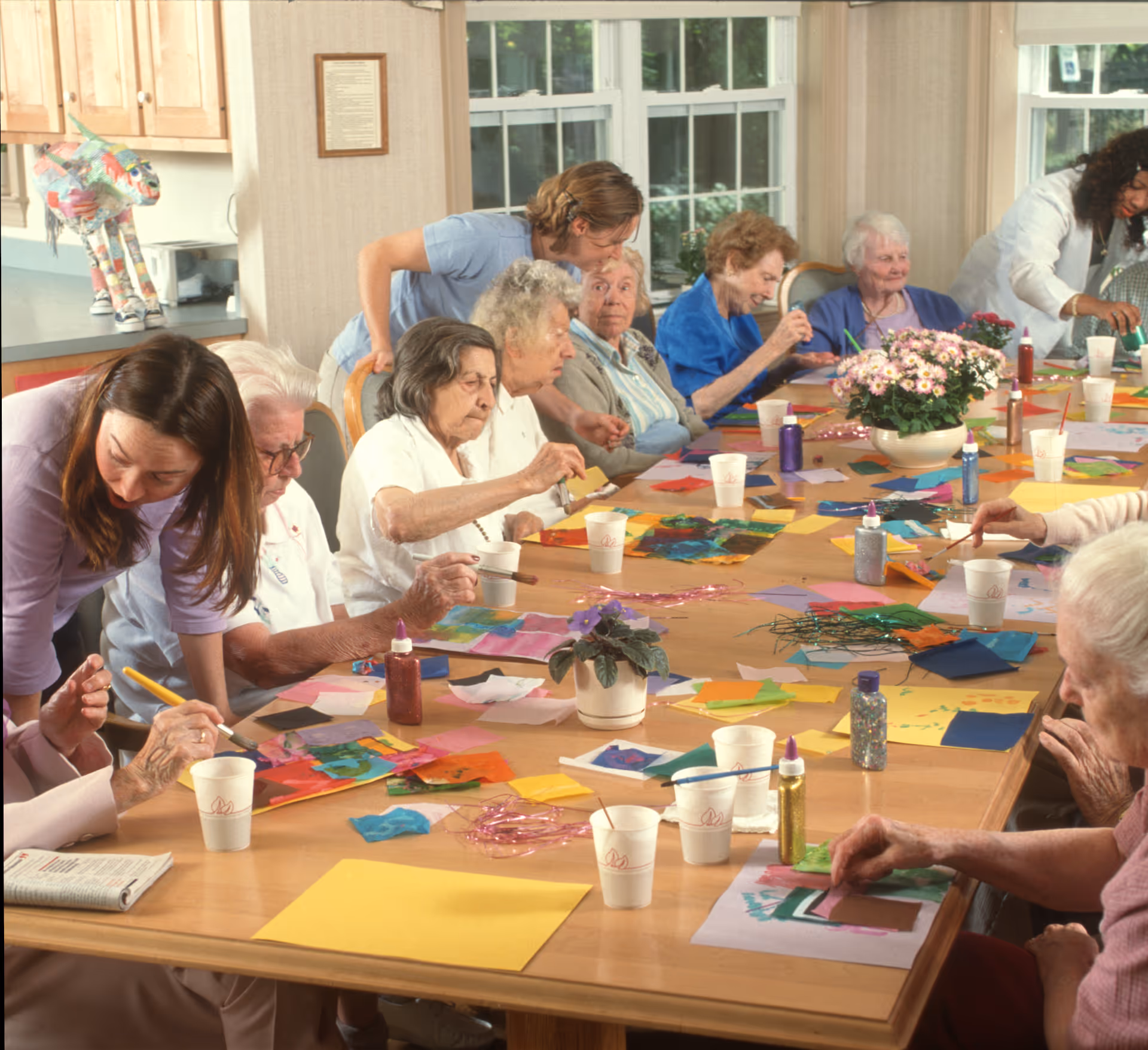 A group of elderly individuals seated around a large table engaged in arts and crafts activities, with colorful paper, glue, and paint supplies spread out. Two caregivers are assisting and interacting with the seniors in a bright room with large windows and potted plants on the table.