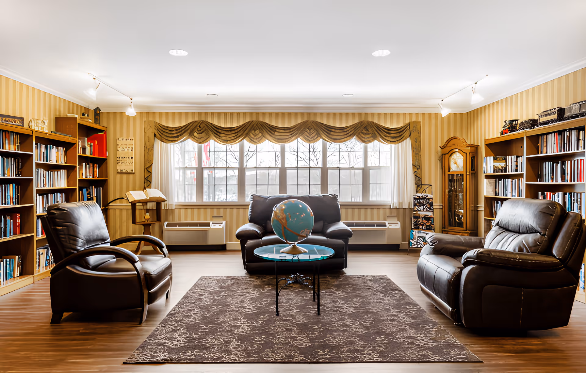 A cozy senior living common room with three dark leather armchairs arranged around a small round glass table holding a globe. The room features large windows with decorative valances, bookshelves filled with books on both sides, a grandfather clock, and a patterned area rug on a wooden floor.